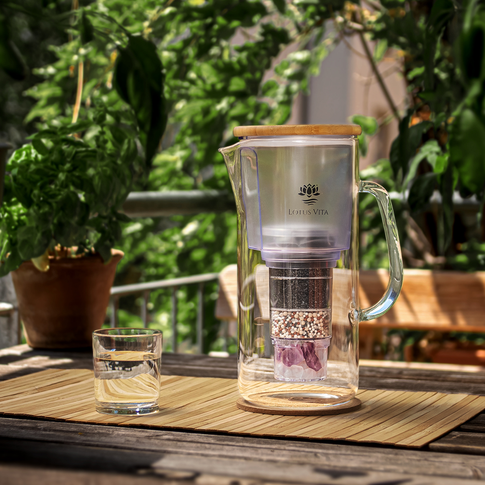 Eine Glas-Filterkaraffe mit Bambusdeckel und einem Glas gefilterten Wassers steht auf einem Holztisch auf einem sonnigen Balkon.
