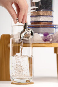 Eine Hand bedient den Zapfhahn eines Glas-Wasserfilters mit Holzständer und füllt frisches, gefiltertes Wasser in ein Trinkglas.
