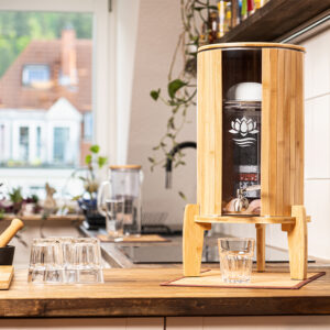 Ein Wasserfilter-Spender aus Glas und hellem Bambus steht auf einer Küchentheke aus Holz, daneben ein frisch gefülltes Glas Wasser.