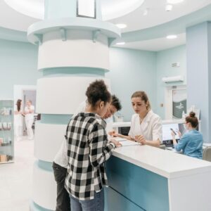 Zwei junge Leute lassen sich am Empfangstresen einer modernen, hellen Arztpraxis von einer medizinischen Fachangestellten beraten.