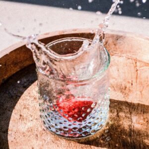 Ein Glas mit frischem, klarem Wasser auf einem Holztablett, in das eine rote Beere fällt und das Wasser spritzen lässt.