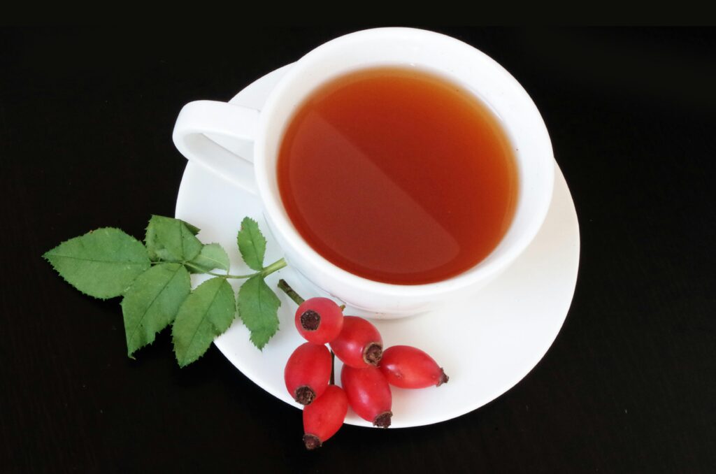 Eine weiße Tasse Hagebuttentee mit frischen Hagebutten und grünen Blättern auf einem schwarzen Untergrund.