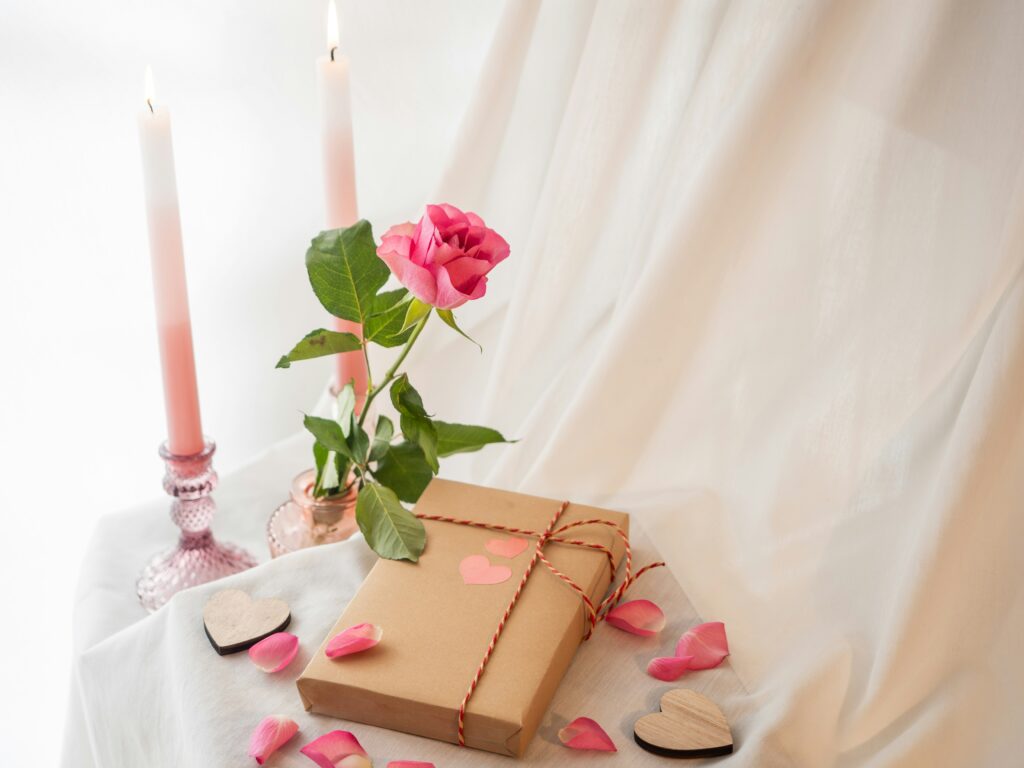 Eine romantische Szene mit einem in Packpapier gewickelten Geschenk, einer einzelnen rosa Rose in einer Vase und zwei brennenden rosa Kerzen auf einem weißen Tuch.