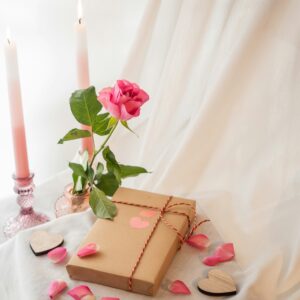 Eine romantische Szene mit einem in Packpapier gewickelten Geschenk, einer einzelnen rosa Rose in einer Vase und zwei brennenden rosa Kerzen auf einem weißen Tuch.