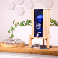 Ein Wasserfilter mit Bambusgehäuse und blauem Glasbehälter steht auf einer Holztheke, daneben ein Tablett mit Gläsern.